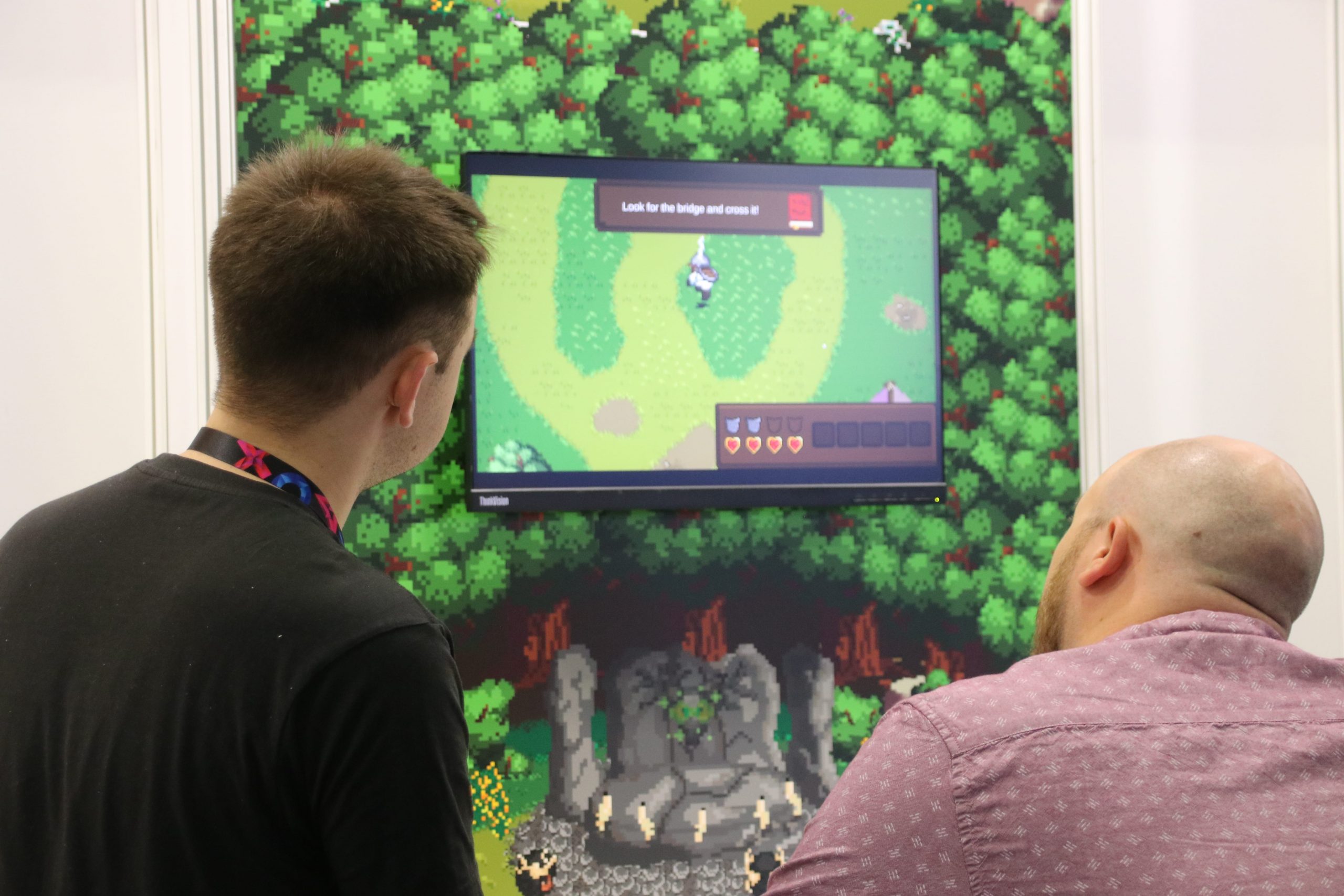 Two males looking at a video game on a TV on a green minecraft style wall